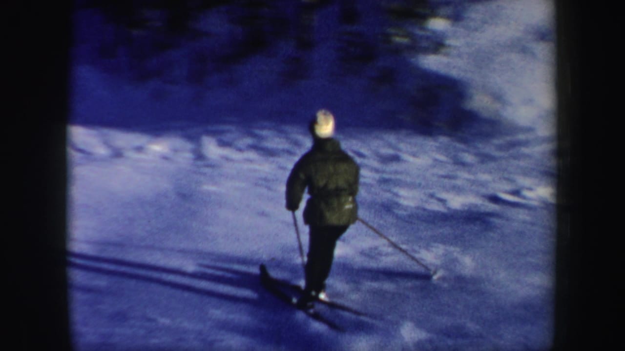 Rear view of the skier in a dark green jacket and white hat gliding over a snowy slope in the 1962 New Hampshire home movie.