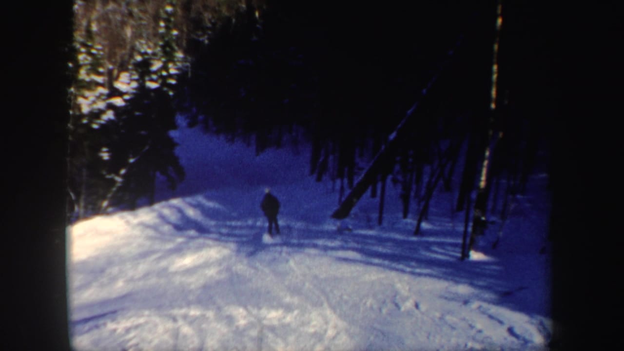 The skier appears smaller in the frame while moving into a darker section of the snowy forest slope in archival 8mm footage.