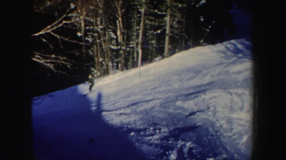 A distant skier crosses a New Hampshire snow slope toward shade, with dark evergreens and bare trees framing the run.