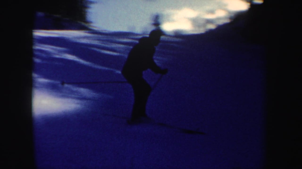 1962 New Hampshire 8mm frame showing a skier in dark clothing on a snowy slope, silhouetted against blue shadows and bright sky.
