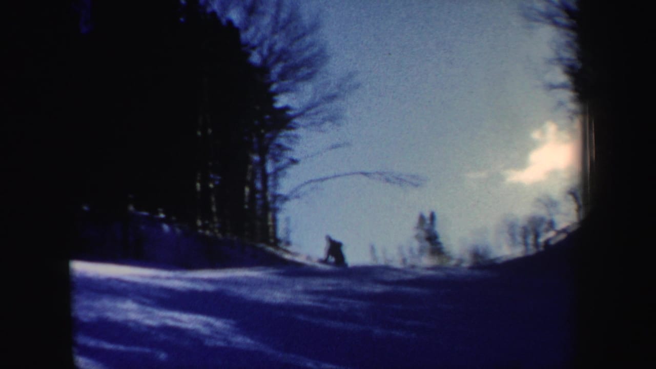Wide 1962 New Hampshire home-movie frame with a distant skier moving across a snowy slope beneath bare trees and pale sky.