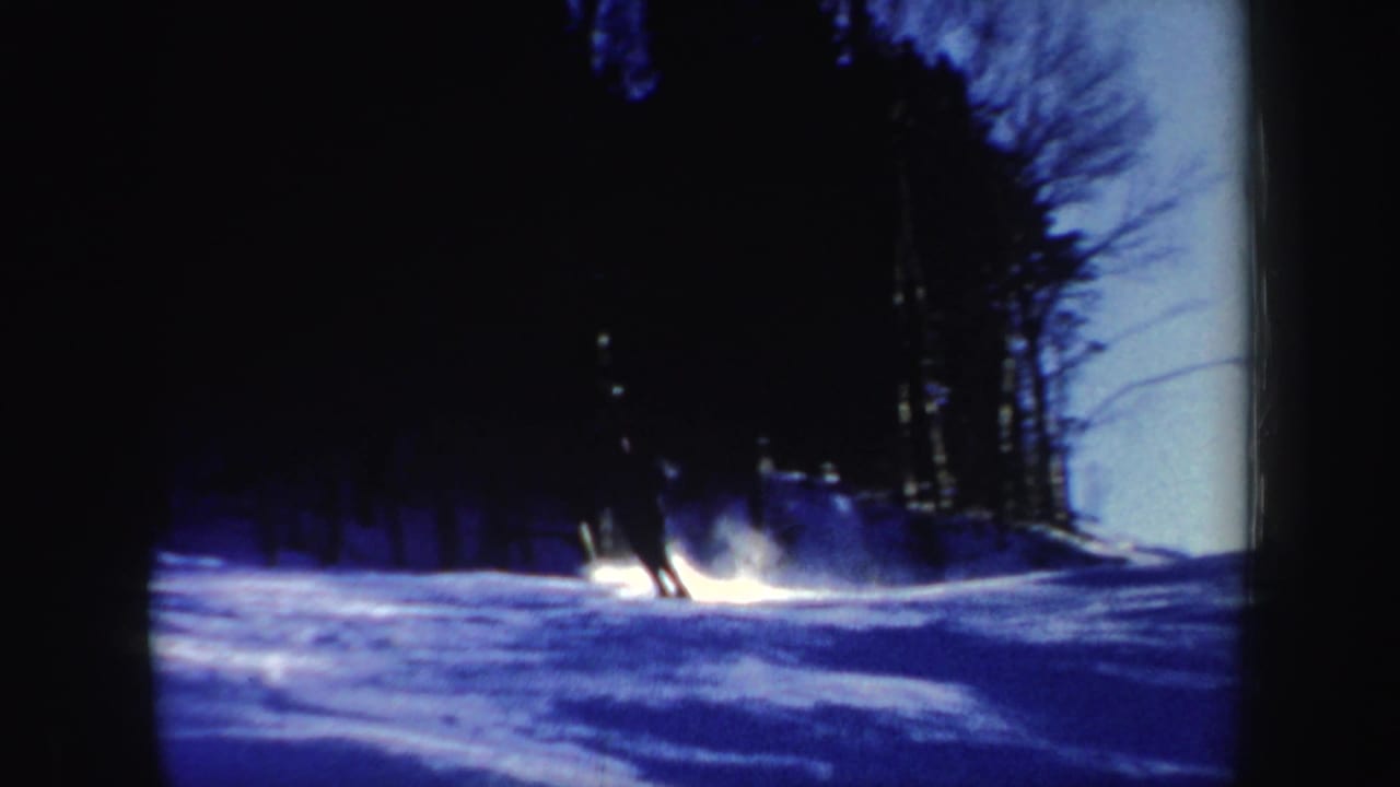 1962 New Hampshire 8mm frame showing a skier turning through blue-shadowed snow near a dark stand of bare trees.