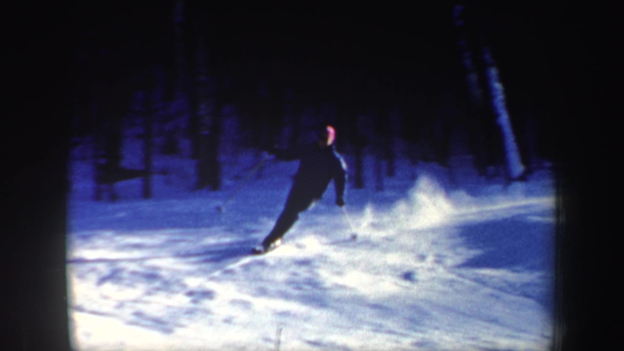 1962 New Hampshire home-movie frame of a skier descending in dark clothing with a red hat visible against blue snow and trees.