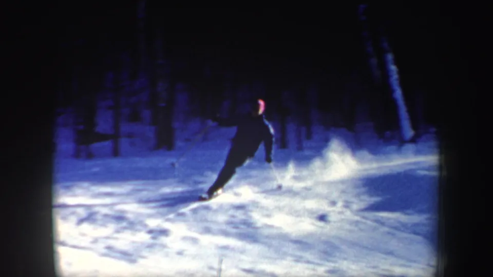 1962 New Hampshire home-movie frame of a skier descending in dark clothing with a red hat visible against blue snow and trees.