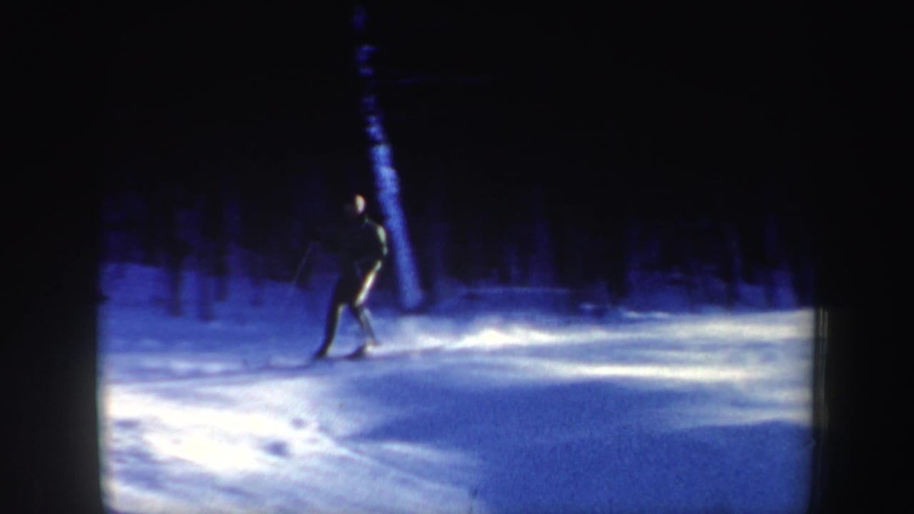 The skier continues downhill across uneven snow, framed by dark trees and blue-tinted 8mm film exposure.