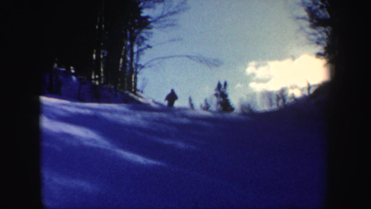 1962 New Hampshire home-movie frame showing a distant skier moving across a blue-cast snowy slope bordered by dark trees.
