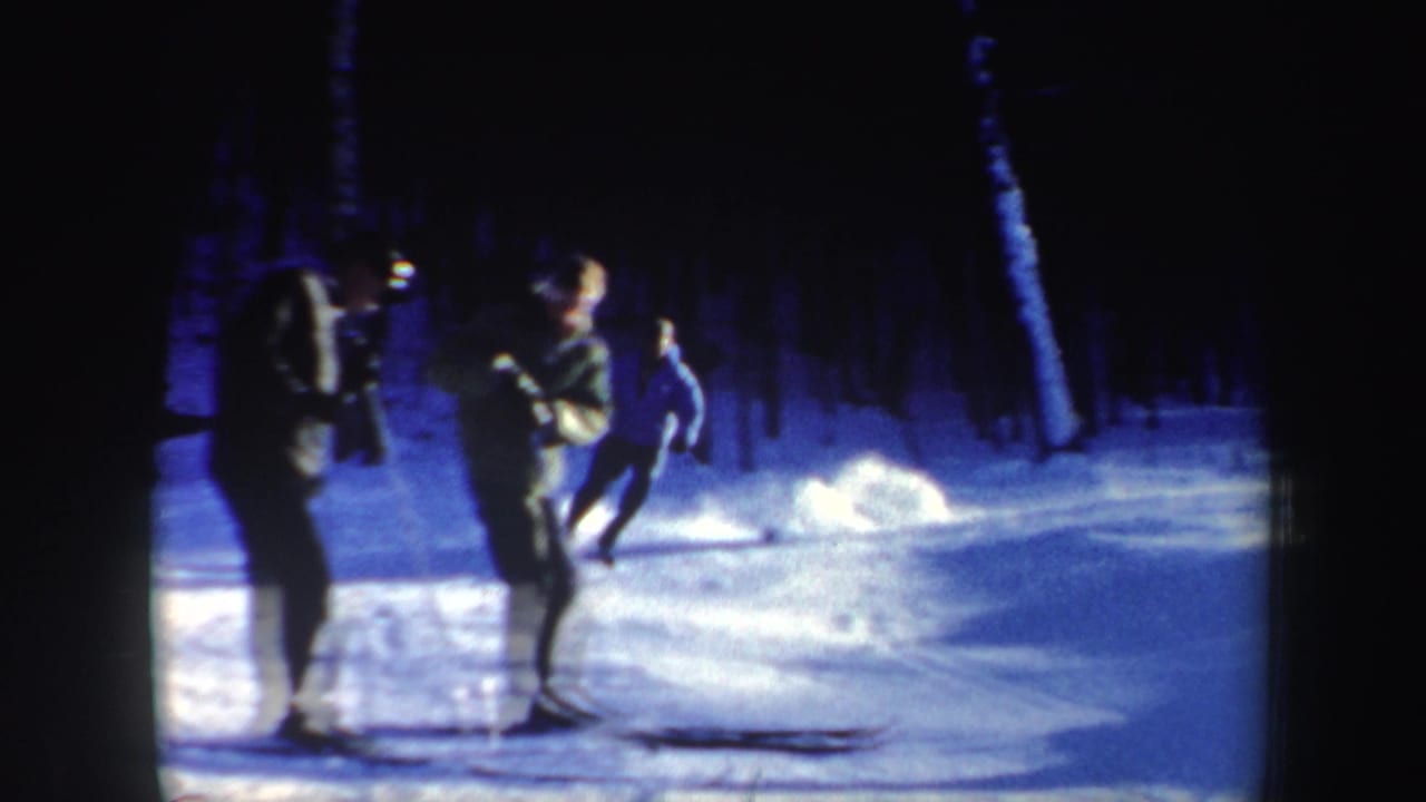 1962 New Hampshire home-movie frame showing three skiers regrouping on a blue-tinted snowy trail near a wooded slope.