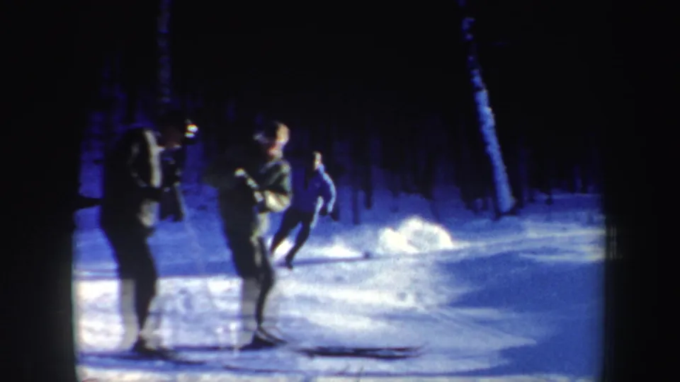 1962 New Hampshire home-movie frame showing three skiers regrouping on a blue-tinted snowy trail near a wooded slope.