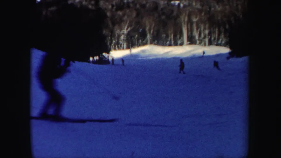 1962 New Hampshire 8mm frame showing distant skiers moving down a gentle snow-covered slope near a wooded edge.
