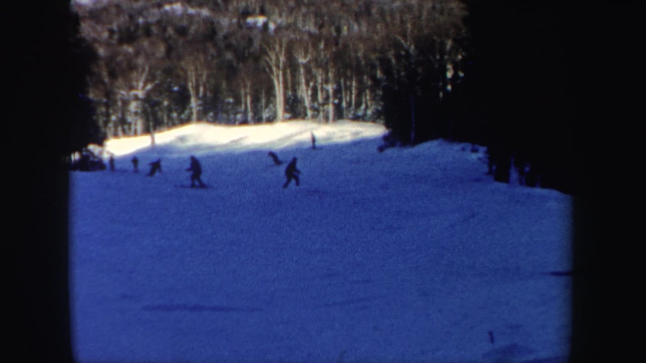 1962 8mm home-movie frame of skiers on a gentle New Hampshire slope, with trees and deep blue snow shadow visible.