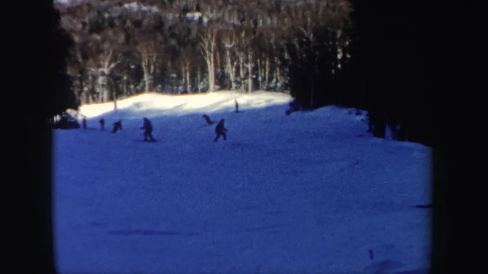 1962 8mm home-movie frame of skiers on a gentle New Hampshire slope, with trees and deep blue snow shadow visible.