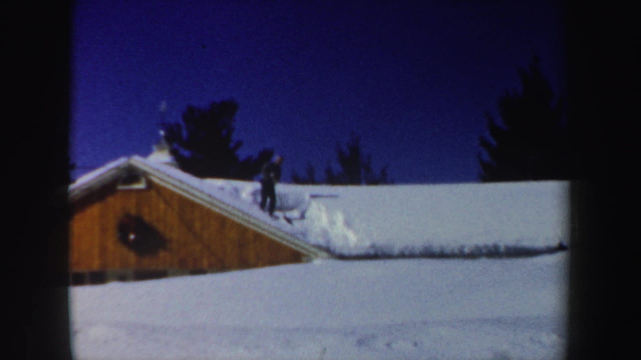 Man stands on a snow-covered house roof beneath a dark blue winter sky.