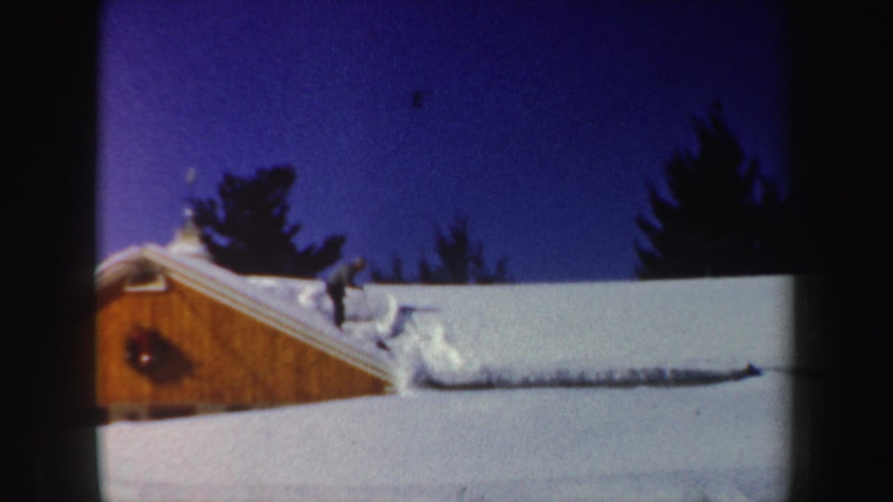 The man continues clearing snow near a drift on the roof, with the wooden house gable and winter sky visible.