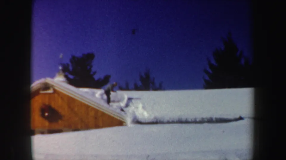 The man continues clearing snow near a drift on the roof, with the wooden house gable and winter sky visible.
