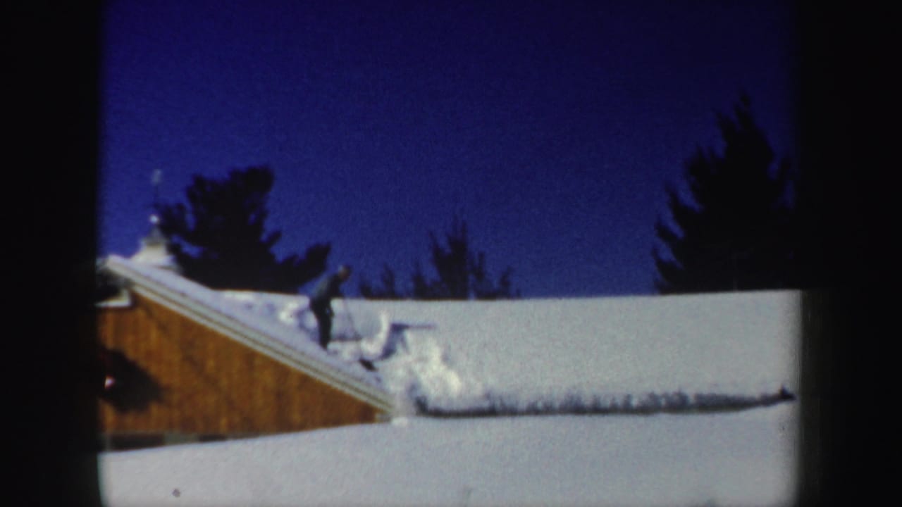 A dark-clothed man pauses or steps on the snow-covered roof beside a mound of cleared snow in the New Hampshire winter scene.
