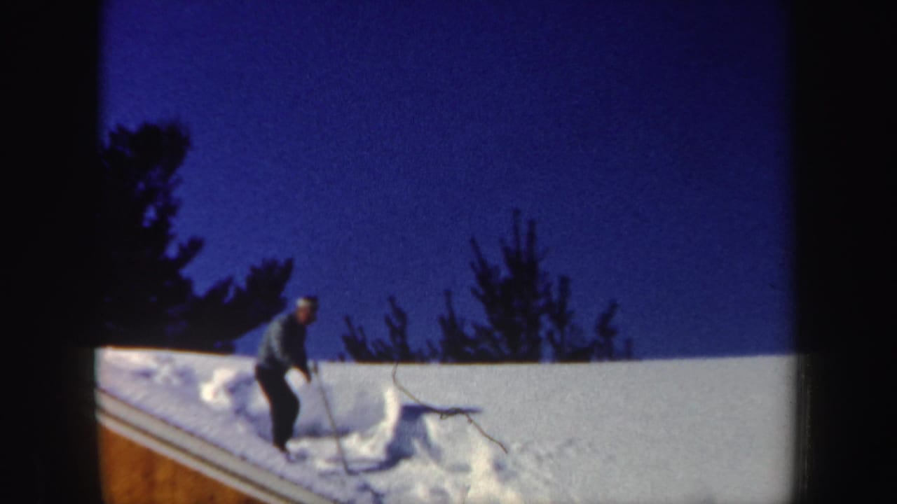 Man standing on a snow-covered sloped roof with a shovel under a blue winter sky.