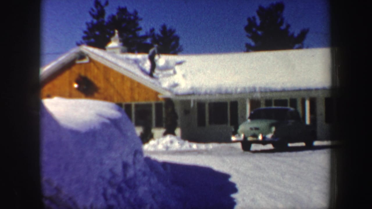 A snowy New Hampshire house with a man clearing the roof and a vintage green car in the driveway, filmed in 1962.