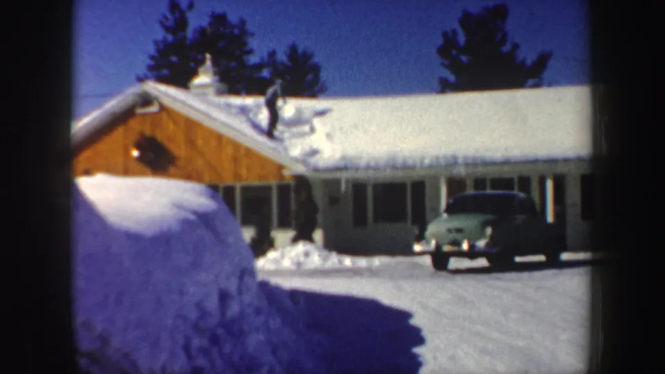 A snowy New Hampshire house with a man clearing the roof and a vintage green car in the driveway, filmed in 1962.