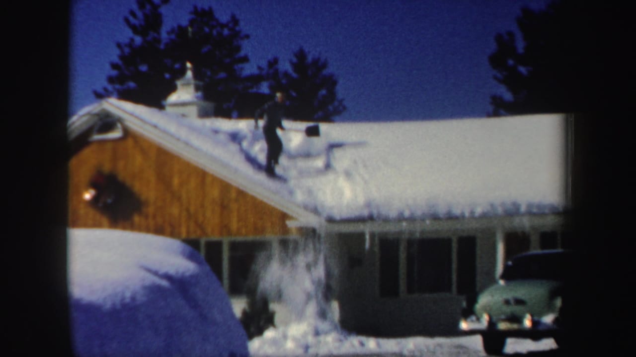 The man pushes snow off the roof edge, sending it down beside the house in this 1962 New Hampshire winter scene.