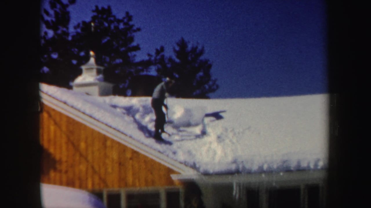 Man shoveling snow on a sloped house roof under a clear sky.