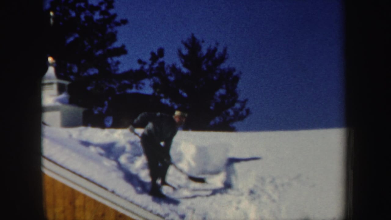 A man in dark winter clothing shovels through deep snow on the roof, with a cleared strip visible beside the wood-sided wall and trees beyond.