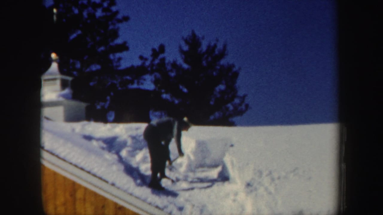 Near the end of the clip, the man keeps shoveling along the sloped roof, surrounded by thick snow, evergreen silhouettes, and clear blue sky.