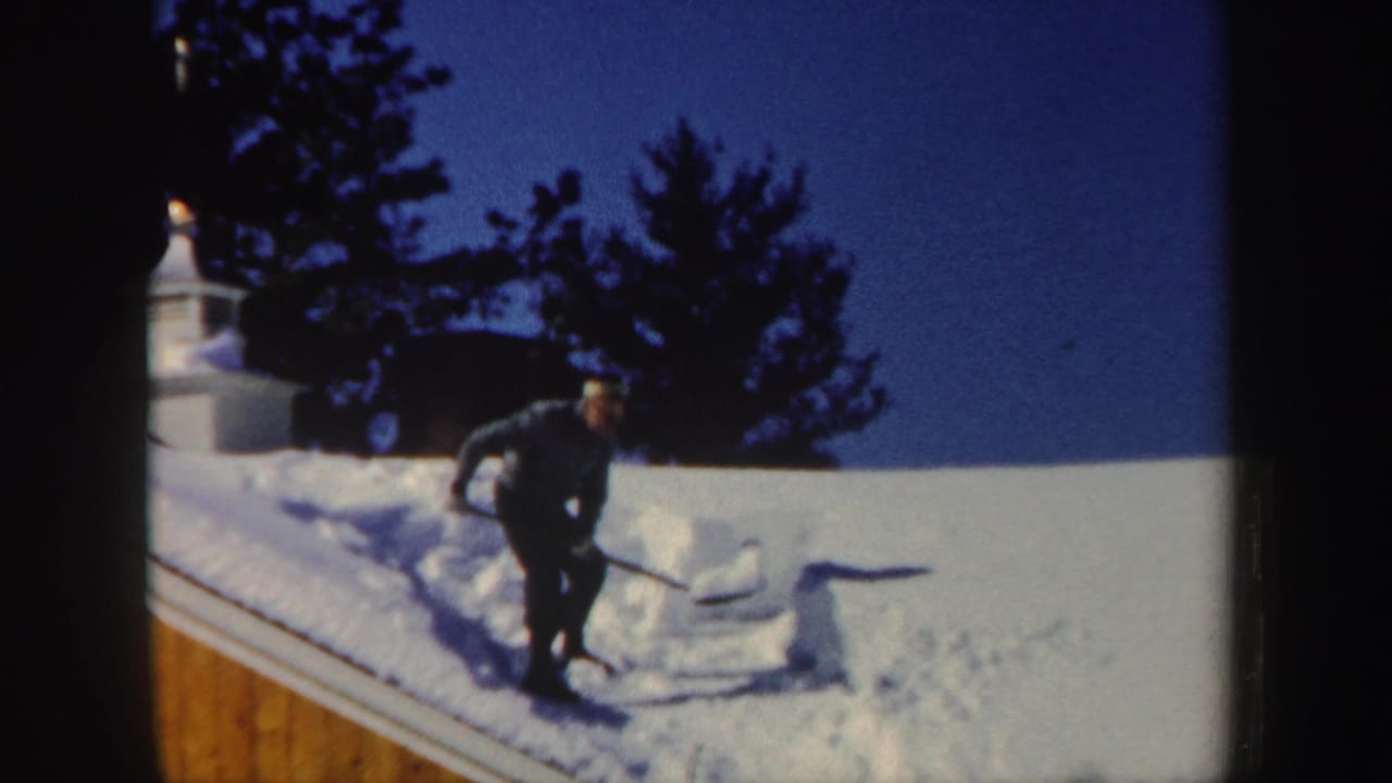 Man shoveling snow on a roof beside a wooden house wall.