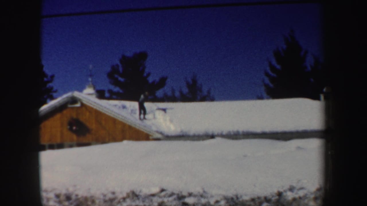 Wide 8mm frame of a man on a snow-covered roof in New Hampshire, with evergreens and a deep blue winter sky behind him.