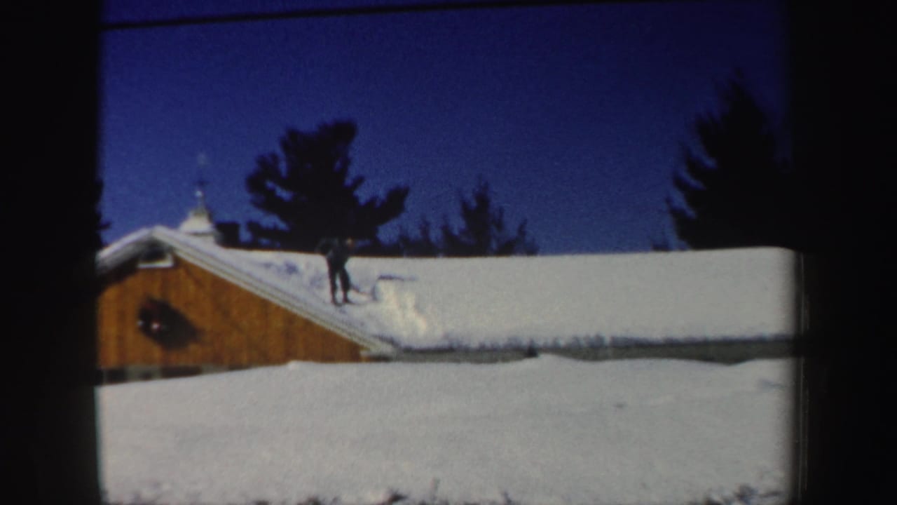 Distant figure in winter clothing works with a shovel on a snow-covered New Hampshire roof in 1962 home-movie footage.