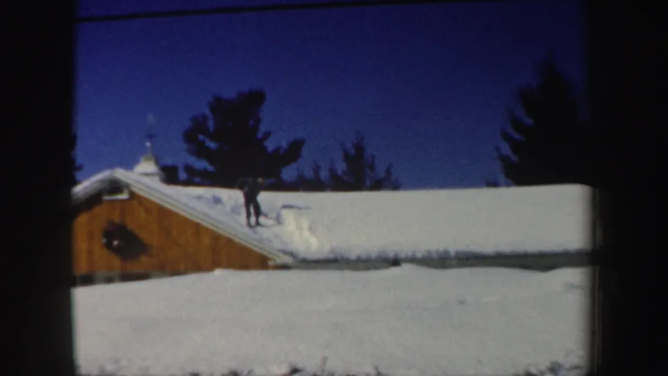 Distant figure in winter clothing works with a shovel on a snow-covered New Hampshire roof in 1962 home-movie footage.