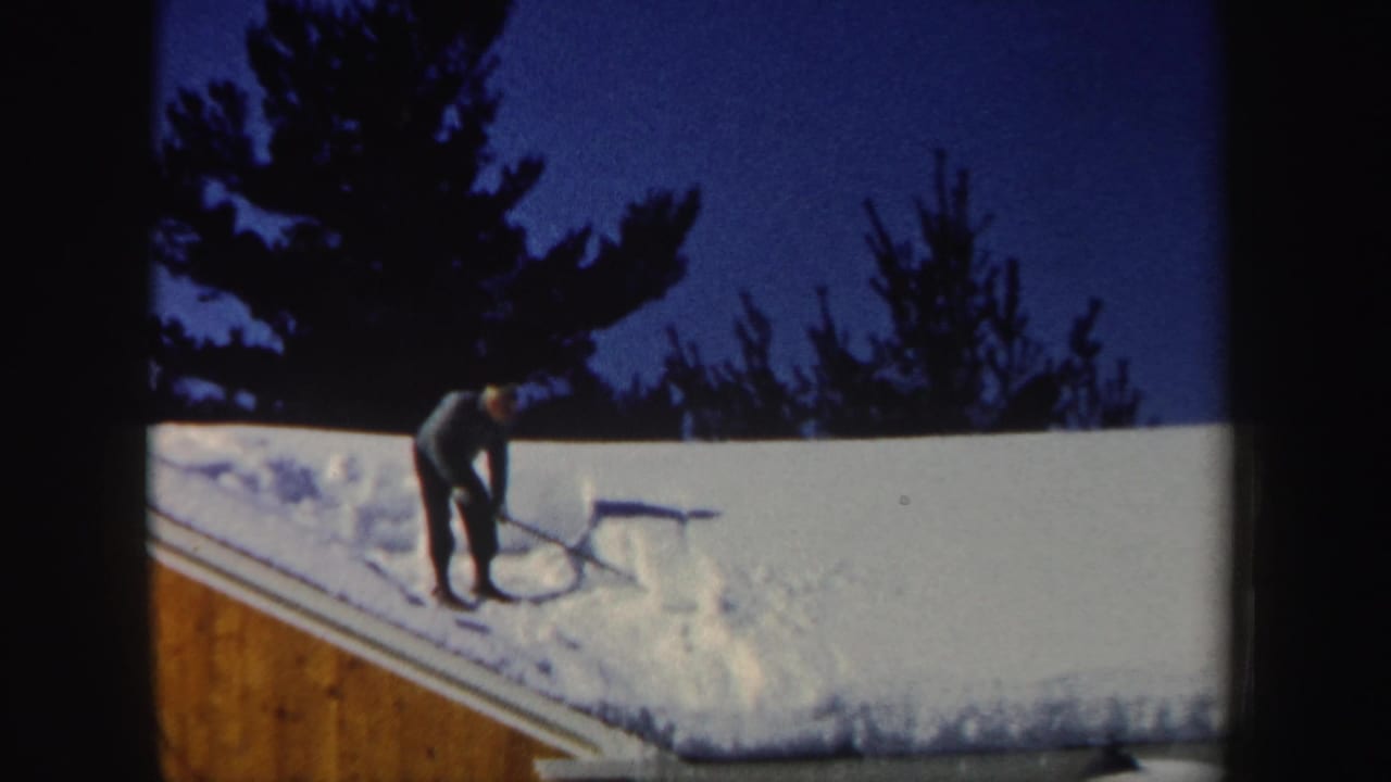 Closer 8mm view of a bundled person shoveling heavy snow from a house roof in New Hampshire.