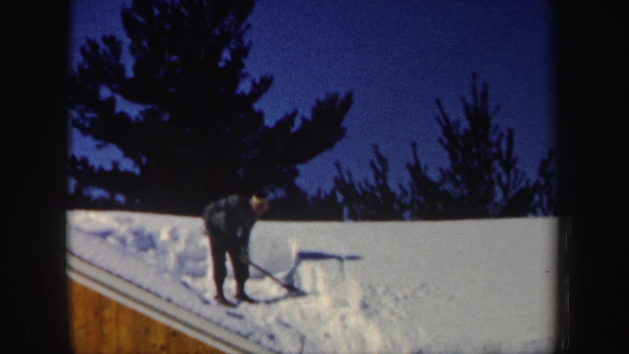 A person in dark winter clothing shovels roof snow in a 1962 New Hampshire home-movie frame.