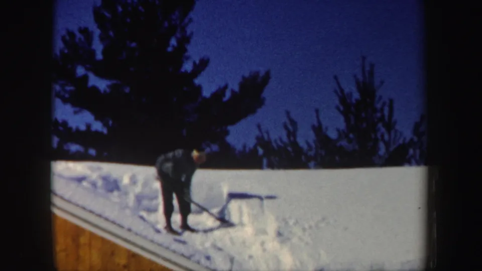 A person in dark winter clothing shovels roof snow in a 1962 New Hampshire home-movie frame.