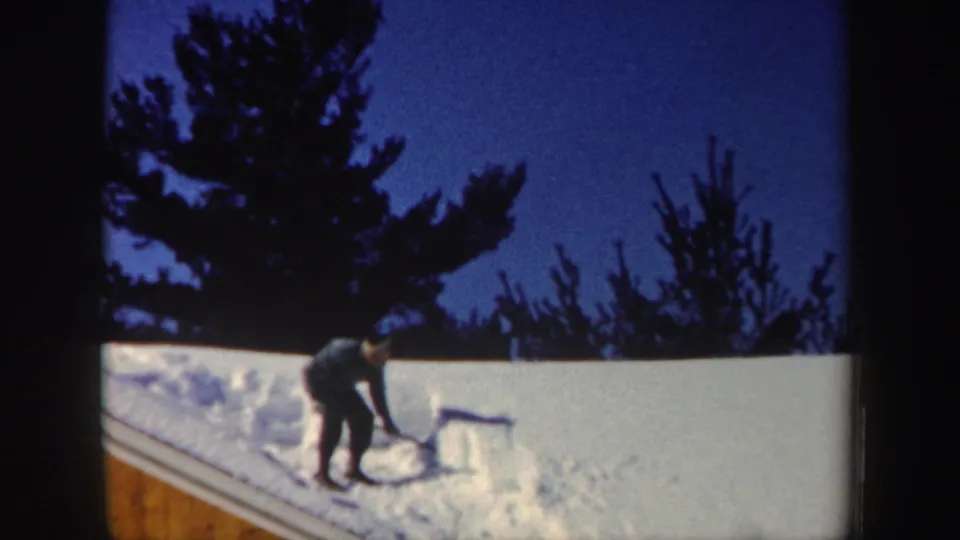 In metadata-supported New Hampshire, 1962, a man in winter clothing shovels snow from a roof under a deep blue twilight sky.