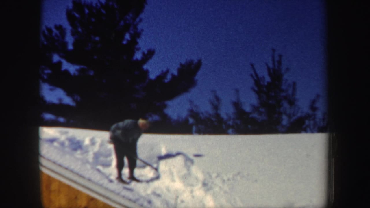 A man works a shovel into packed roof snow, with dark evergreen silhouettes and blue twilight sky behind him.