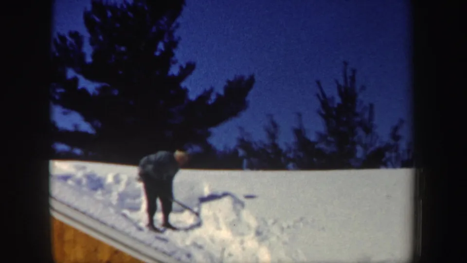 A man works a shovel into packed roof snow, with dark evergreen silhouettes and blue twilight sky behind him.