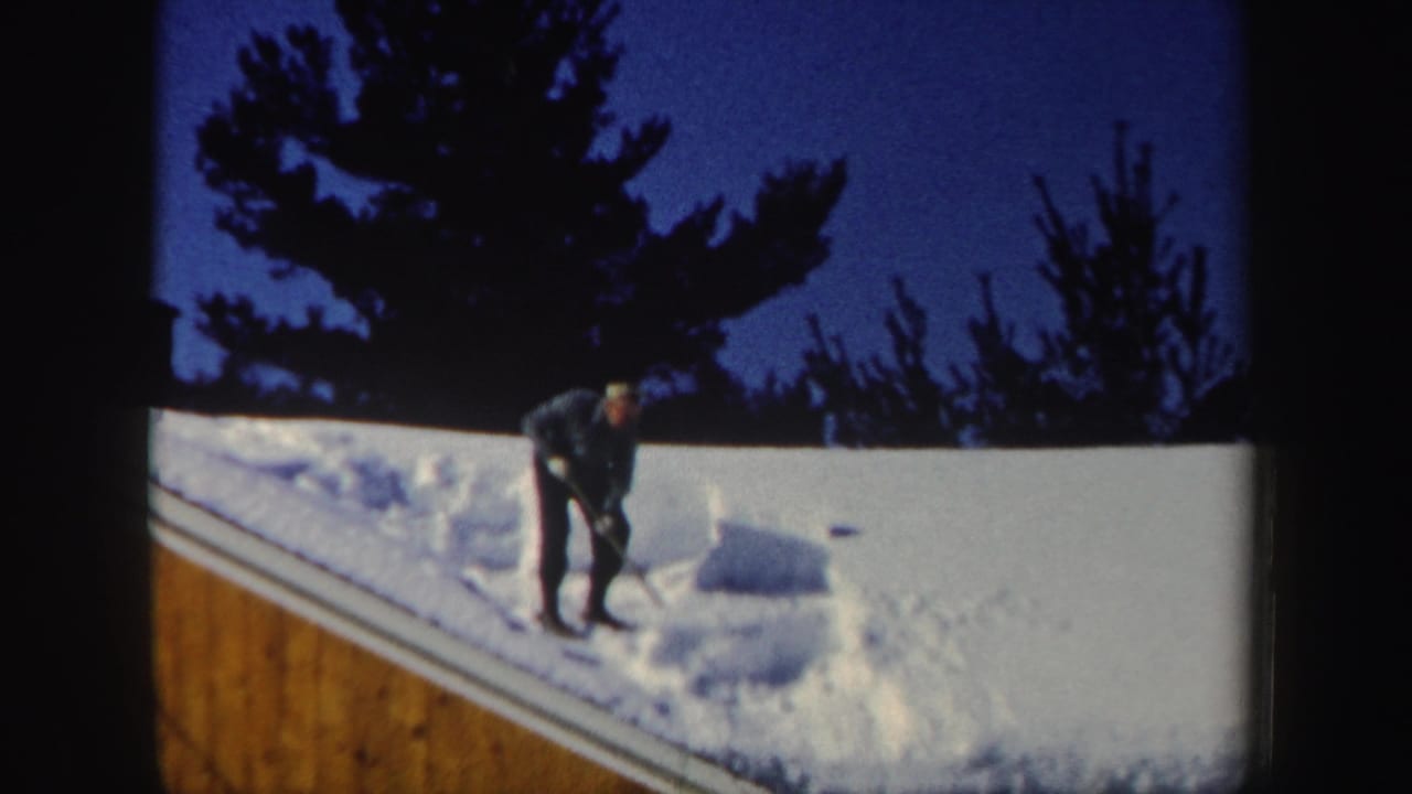 The man stands on the sloped roof after moving snow aside, leaving a cleared patch near the roof edge in the 1962 New Hampshire clip.