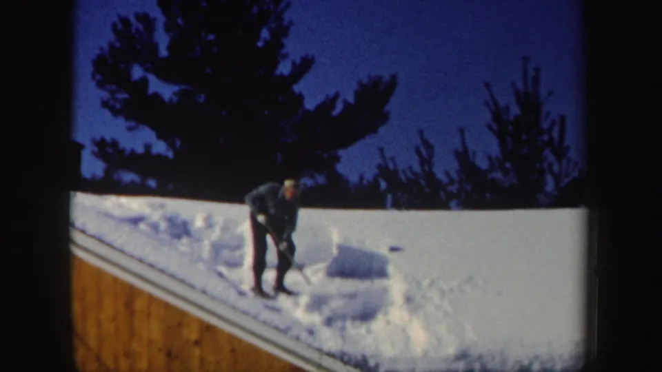 The man stands on the sloped roof after moving snow aside, leaving a cleared patch near the roof edge in the 1962 New Hampshire clip.