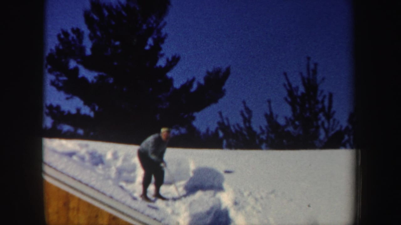 A man pauses or resets his stance with a shovel on a snow-covered roof, framed by dark trees and twilight sky.