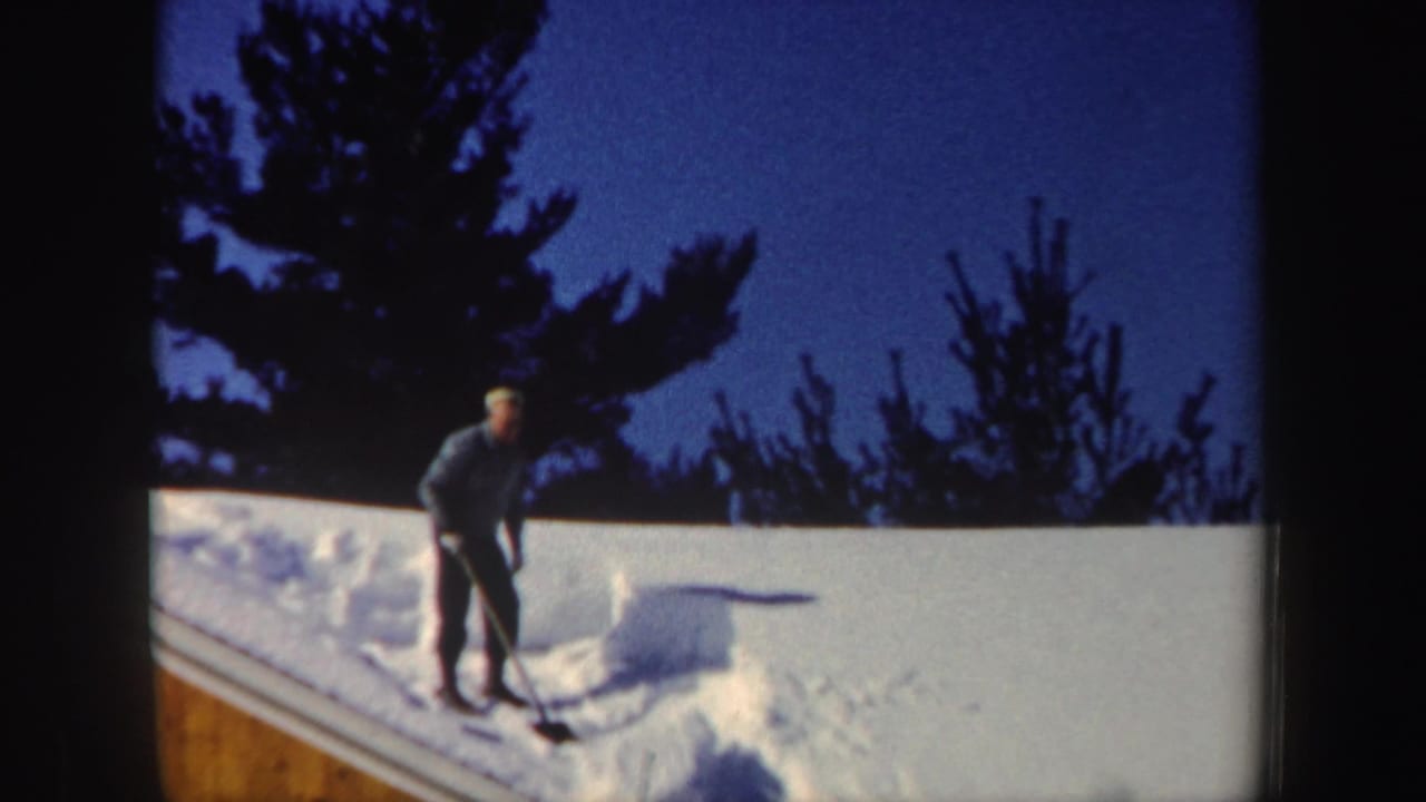 Man standing on a snow-covered roof with a shovel near dark trees.
