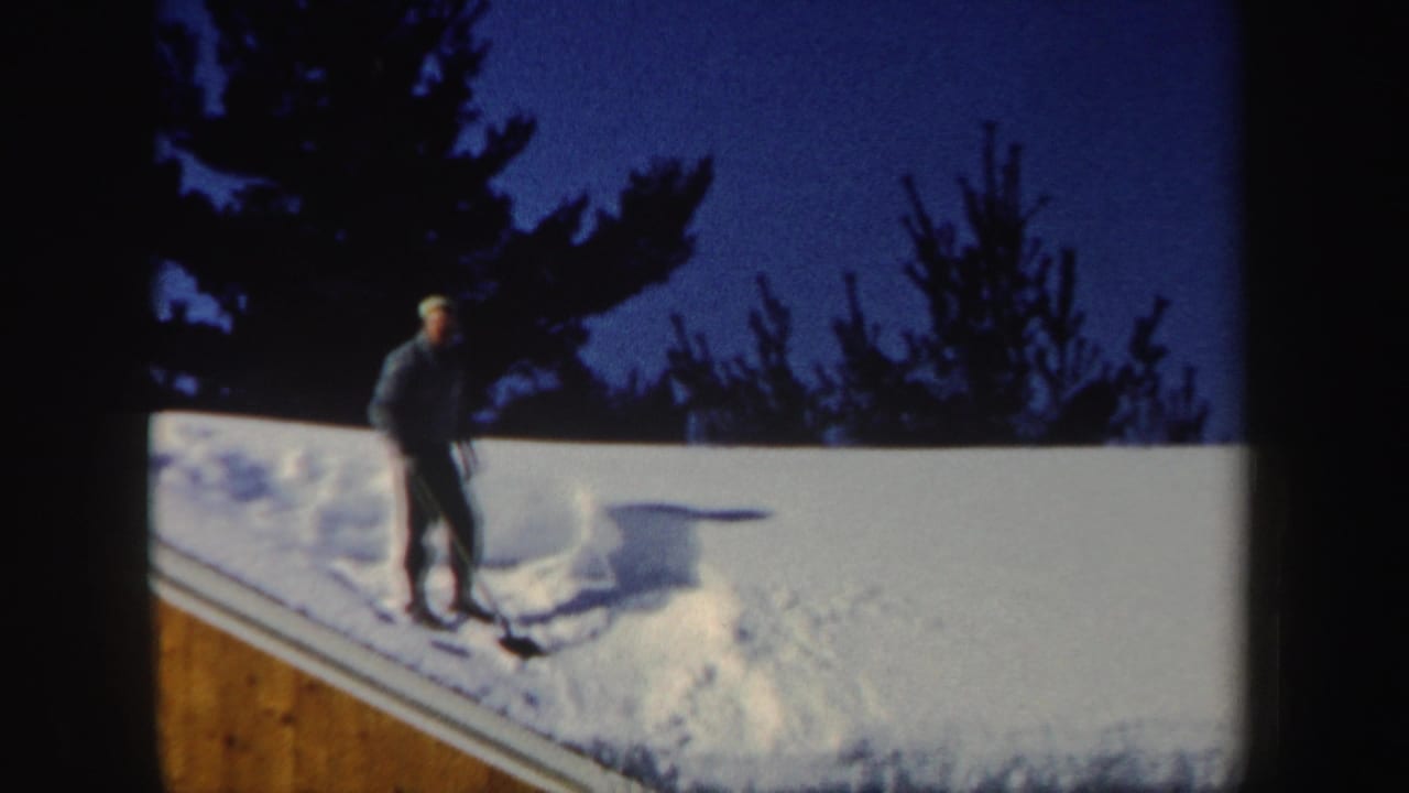 A man in a dark coat and hat shovels deep roof snow in a snowy New Hampshire winter scene from 1962.