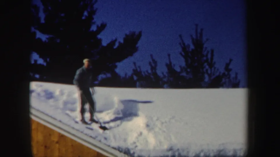 A man in a dark coat and hat shovels deep roof snow in a snowy New Hampshire winter scene from 1962.