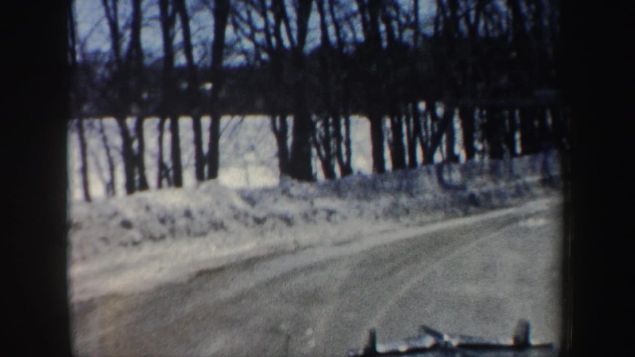 Snowy two-lane road seen through a vehicle windshield, with bare trees and snowbanks along the shoulder.