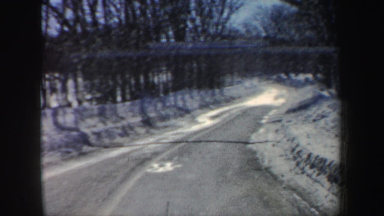 Windshield view of a curving snowy New Hampshire road in 1962, with plowed shoulders, bare trees, and winter light on the pavement.