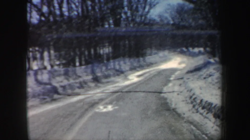 Windshield view of a curving snowy New Hampshire road in 1962, with plowed shoulders, bare trees, and winter light on the pavement.