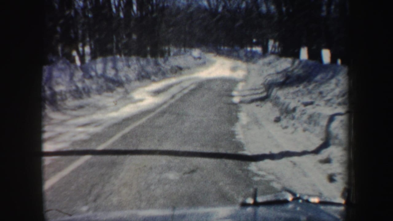 1962 New Hampshire home-movie view from a car approaching a snow-lined road, with bare trees and the vehicle hood visible at the bottom.