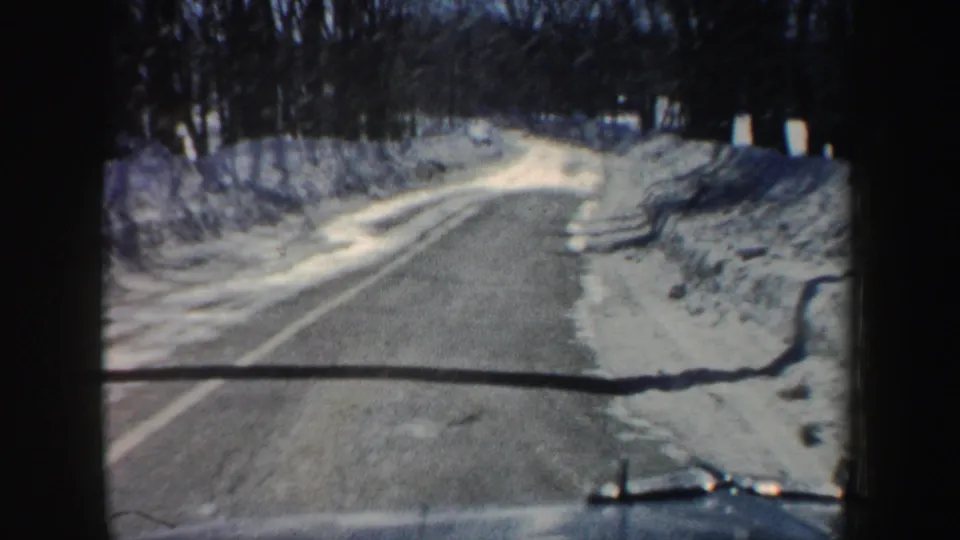 1962 New Hampshire home-movie view from a car approaching a snow-lined road, with bare trees and the vehicle hood visible at the bottom.