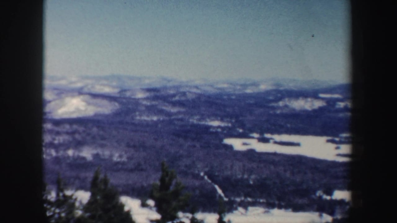 Elevated New Hampshire winter landscape on 8mm film, showing frozen lake areas, dark wooded terrain, distant snowy ridges, and scan frame edges.