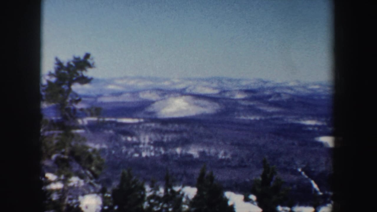 1962 home-movie frame of a New Hampshire winter overlook with evergreen foregrounds, snowy hills, wooded slopes, and a pale track or stream below.