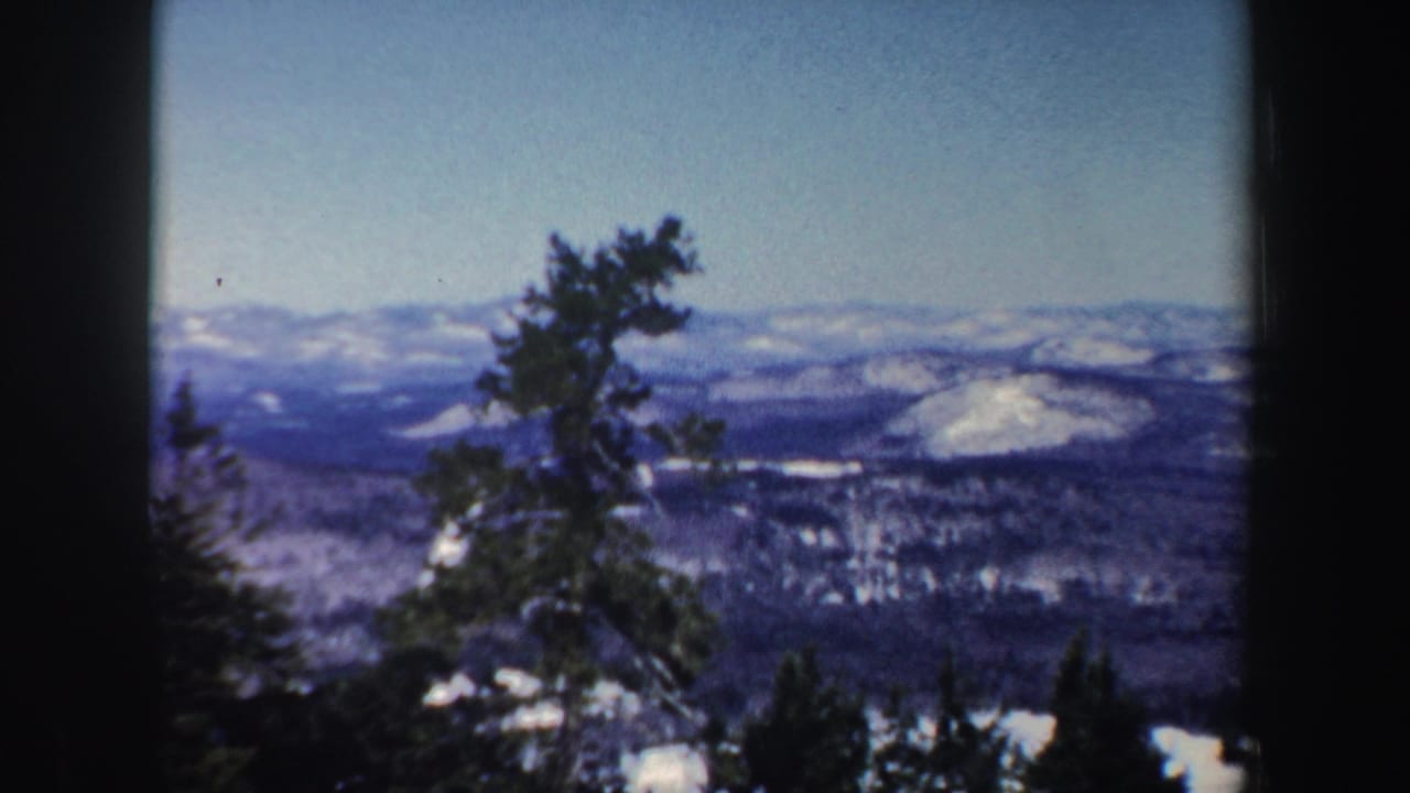 New Hampshire winter landscape on 8mm film with evergreen branches in the foreground, snowy rounded hills, dark forest patches, and a pale sky.
